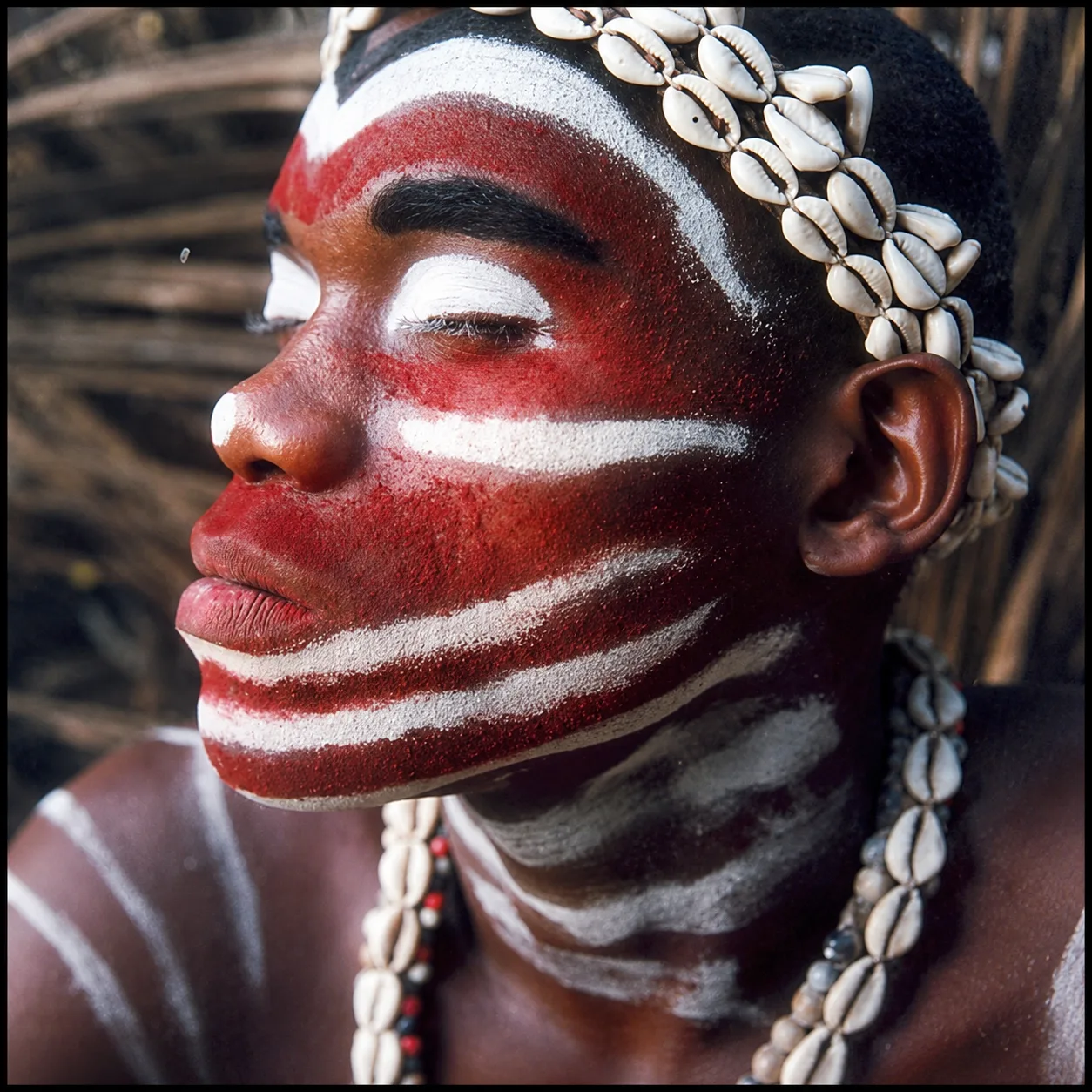 Close-up portrait of an initiate with face painted red and white, adorned with cowries, a serene gaze.