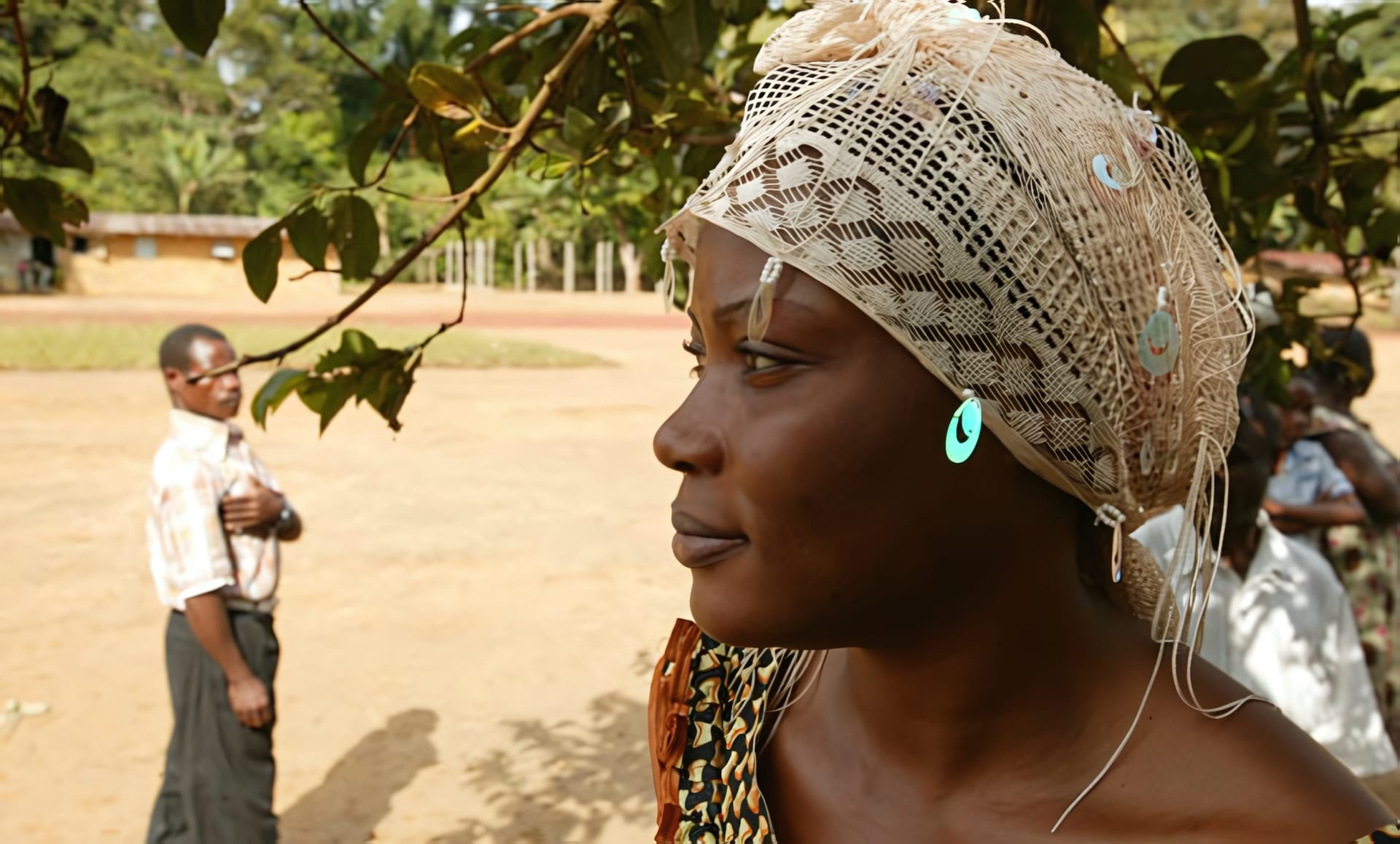 Femme gabonaise de profil, coiffe de dentelle claire, le regard tourné vers la lumière, un homme en retrait sur la place du village
